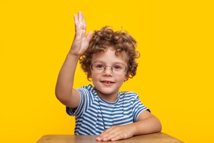 Cute young smart boy in glasses looking at camera with hand up.