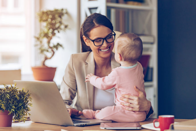 Working together is so fun! Cheerful young beautiful businesswoman looking at her baby girl with smile while sitting at her working place