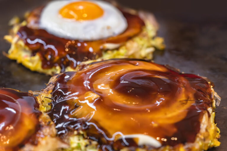 Close-up on a fried egg and an okonomiyaki Japanese omelet prepared with shredded cabbage, cooked on both sides on a hot plate and brushed over in circle with oyster and worcestershire sauce.