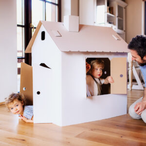 Two happy toddler children with father playing with paper house indoors at home.