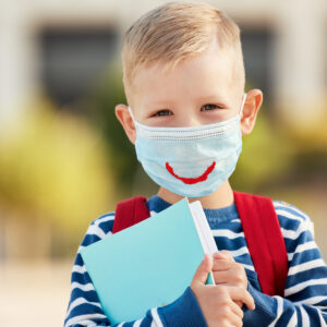 Optimistic smart first grade schoolboy in casual wear and eyeglasses wearing protective mask with drawn smile holding copybooks and looking at camera against blurred urban background